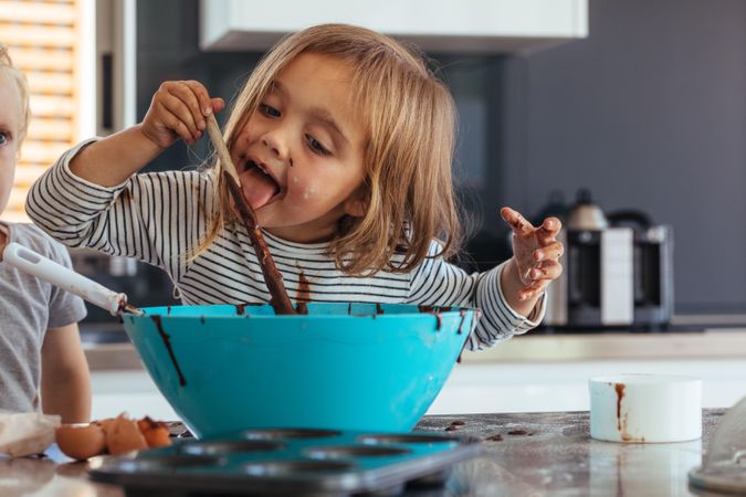 Little girl licking spoon while mixing batter for baking in kitchen  and her brother standing by