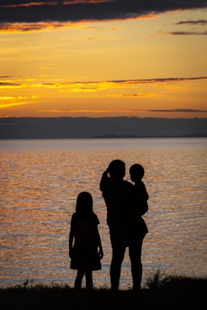 Silhouette of mother holding a baby standing with daughter by waterside at sunset