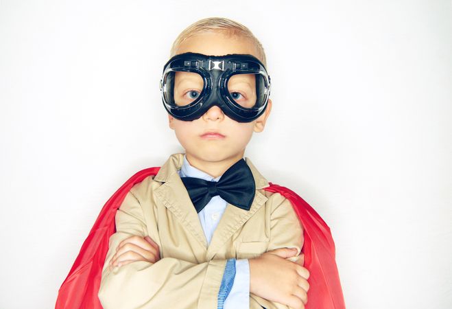 Serious blond boy wearing airplane goggles and cape with his arms crossed