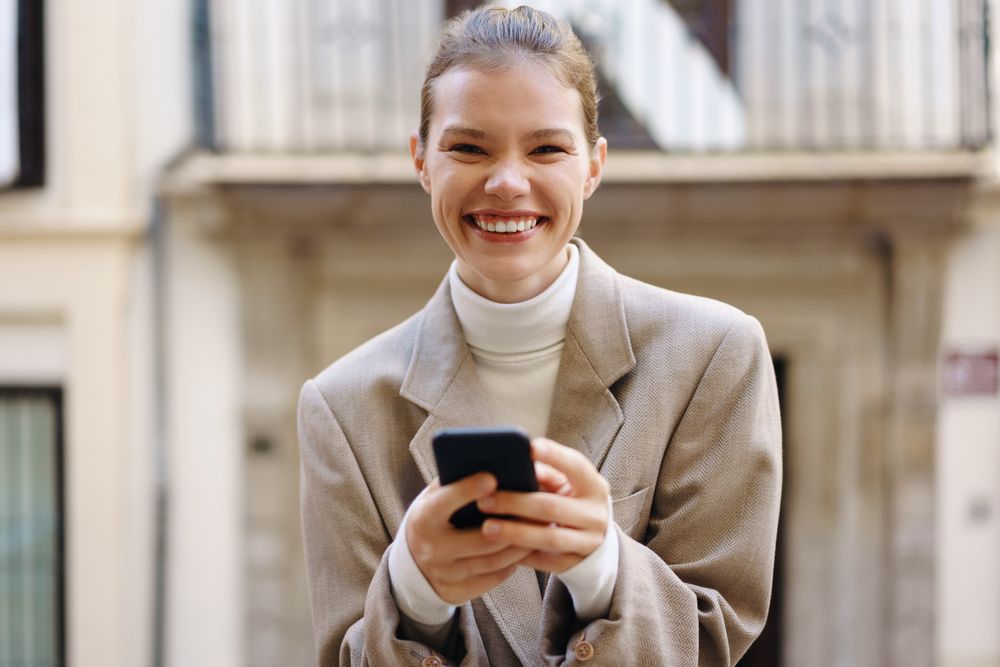 A joyful young woman is smiling brightly while she is texting on her smartphone outdoors