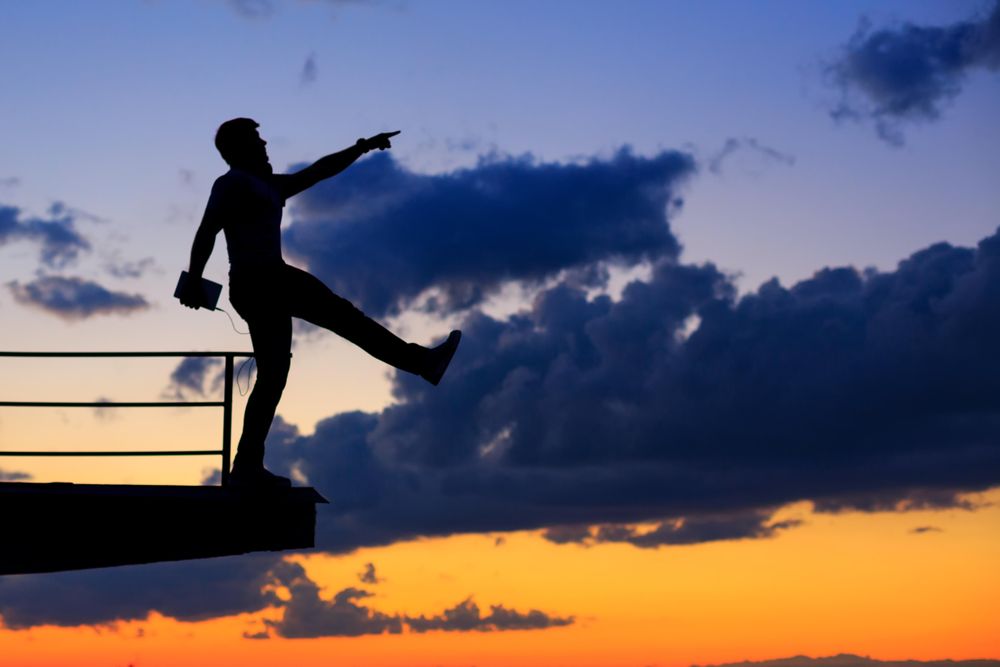 Out line of male walking off ledge and holding tablet against a colorful dusk side, vertical
