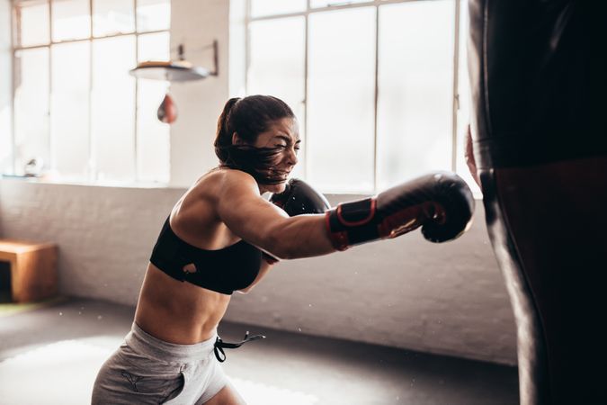 Serious female boxer using a punching bag with gloves inside a boxing ring