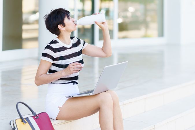 Woman sipping water bottle while working on laptop on steps outside