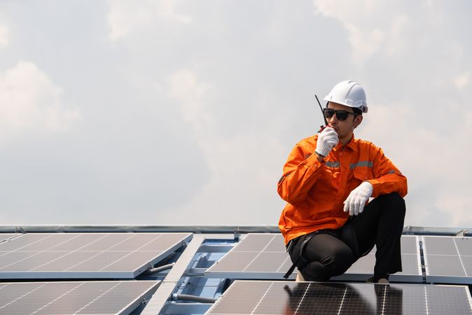 Male engineer in protective work wear talking on walkie talkie with colleague while checking on solar cell or photovoltaic panel installed on the roof of the factory. Industrial renewable energy