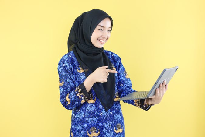 Cheerful Muslim  Woman in Blue Korpri Batik Uniform pointing at Laptop