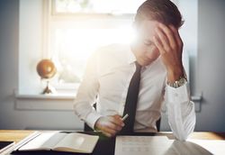 Man sitting at desk reading over documents in a sunny office with his hand to his head
