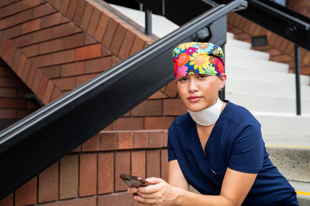 Doctor with head covering and mask around neck sitting outside medical building with smart phone