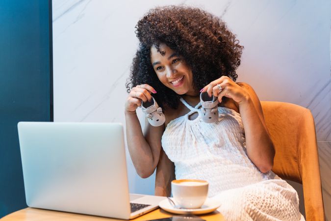 Smiling pregnant Black woman showing baby booties to laptop