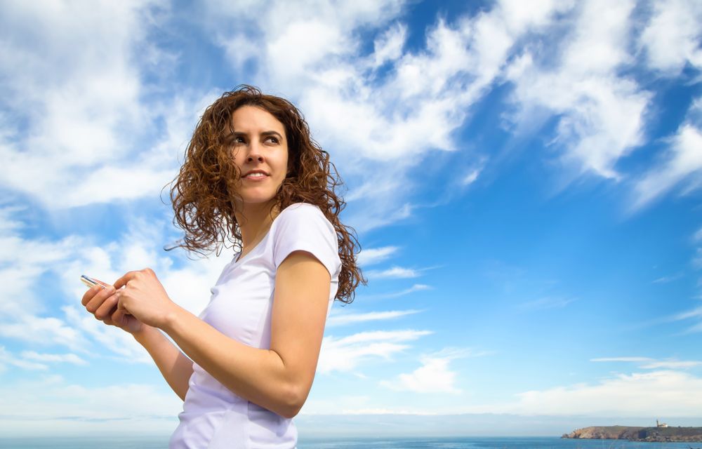 Woman in t-shirt looking around with phone with the coast in background
