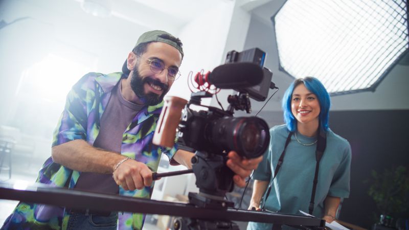 Young Male And Female Filmmakers Collaborate In A Modern Studio, Handling Professional Camera Equipment With Enthusiasm And Expertise, Showcasing Teamwork In Creative Media Production.