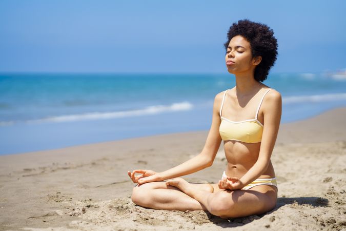 Woman in yellow swimwear sitting cross legged and meditating on beach