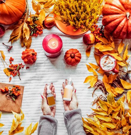Autumn skin care ´beauty products. Women hand holding cosmetic bottles at white background with fall leaves, candles, pumpkins and hot chocolate. Top view with copy space. Beauty lifestyle