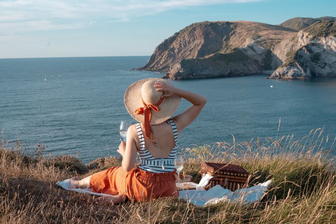 Woman with glass of drink on picnic mat looking at sea and mountains