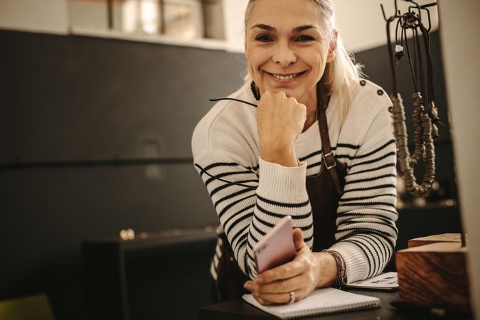 Portrait of smiling female jeweler leaning to desk with phone in hand and looking at camera