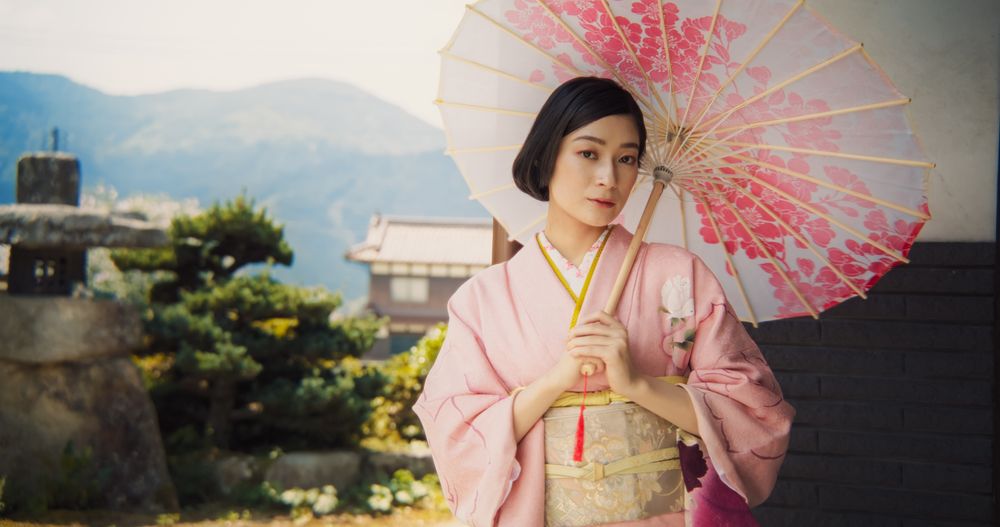 Japanese Female Gently Looking at Camera, Softly Smiling, Wearing a Traditional Pink Kimono with Flower Ornaments and Holding a Stylish Umbrella.