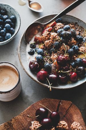 Oat granola yogurt bowl with cherries, blueberries, honey, nuts, coffee, wooden board in foreground