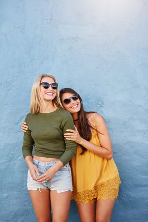 Portrait of two female friends in casuals and sunglasses standing together over blue background