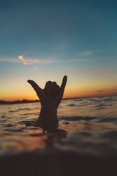 Hand with shaka gesture between water surface and sky at sunset