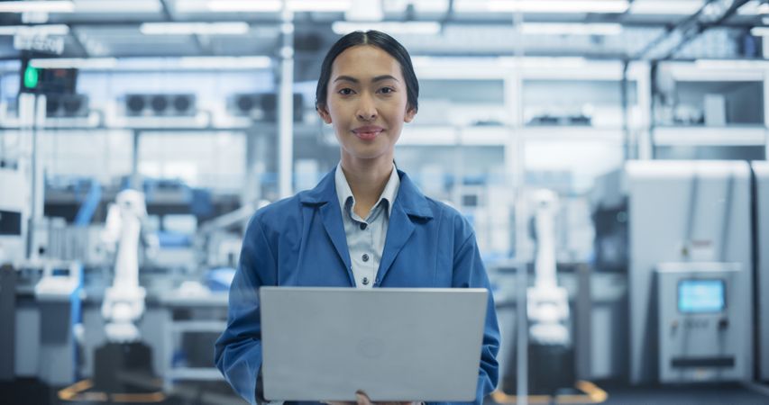 Portrait of Electronics Manufacture Engineer Smiling, Looking at Camera. Young Asian Female Using Laptop Computer for Online Research, Monitoring Industrial Artificial Intelligence Systems