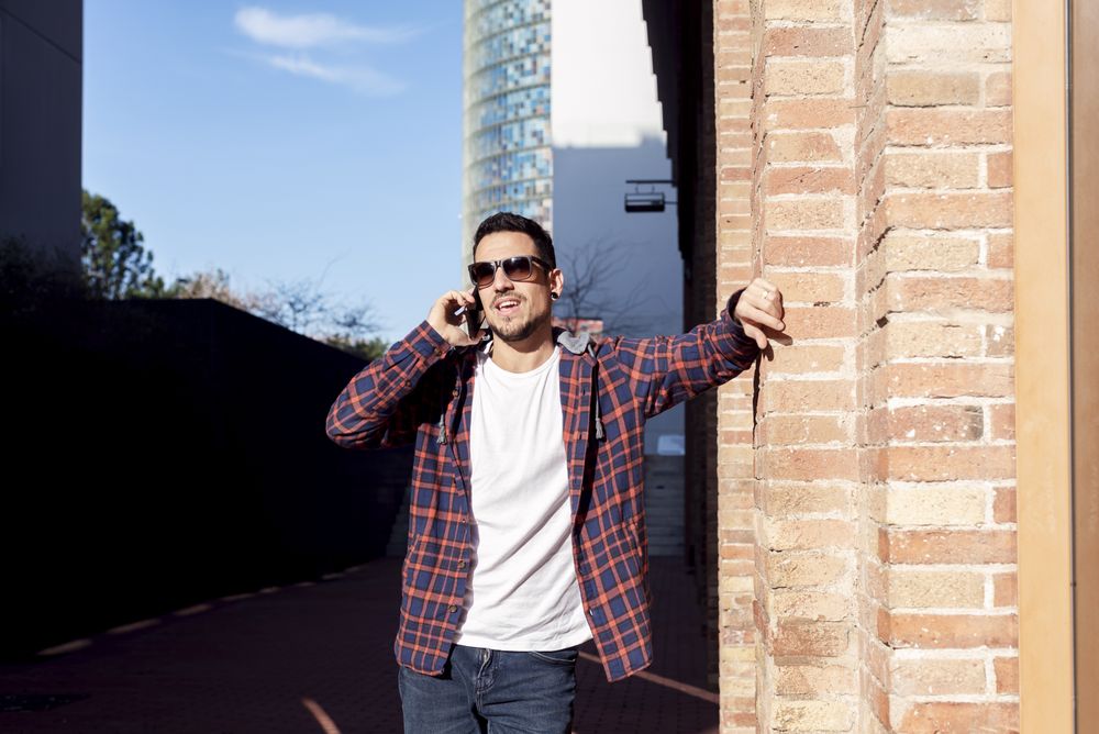 Man standing by brick wall wearing sunglasses and chatting on smartphone outdoors