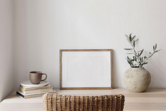 Empty horizontal picture frame mockup. Wooden table, desk with cup of coffee, blurred rattan chair. Vase with olive tree branches, old books. Mediterranean interior, home.
