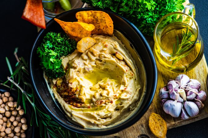 Looking down at creamy hummus dip in bowl on board served with swirl of olive oil and chips