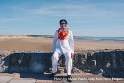 An Elvis impersonator in all white with orange lei sitting on rock wall by the beach 49mJa4