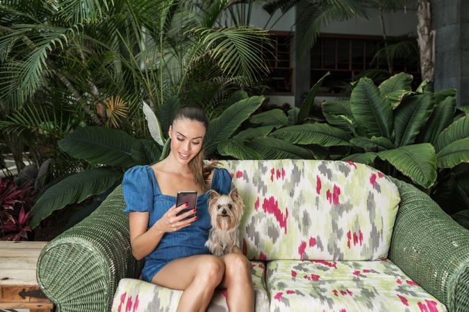 Smiling woman using smartphone sitting beside Yorkshire terrier puppy on floral couch outdoor