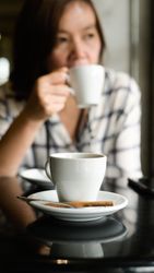 Close up shot A coffee mug is placed on the table, a young woman sits drinking coffee in the background.