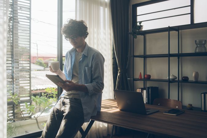Man working from home perched on desk reading tablet with coffee in hand