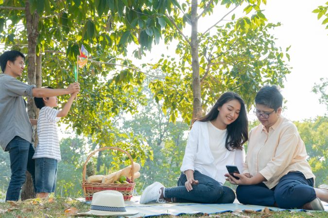 Older woman and her daughter on smartphone in park with son and father playing in background