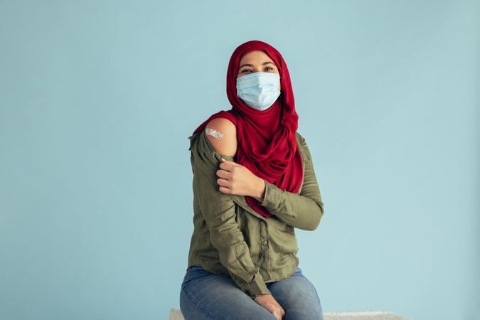 Muslim female after receiving corona virus vaccination sitting against blue background