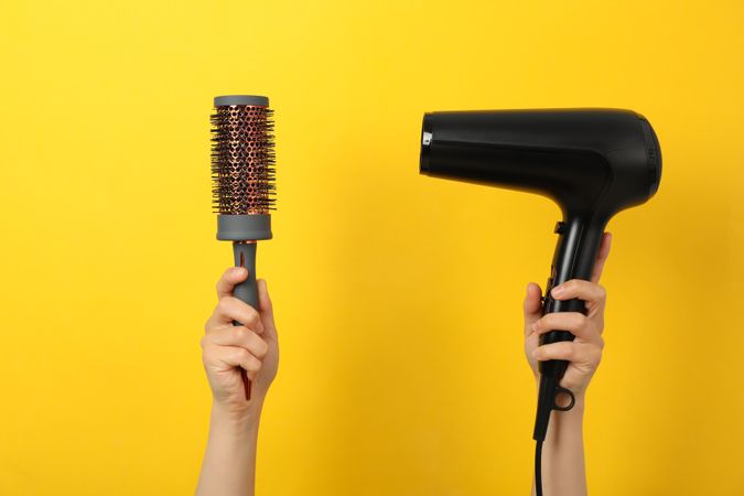 Female hands hold hair dryer and hair brush on yellow background