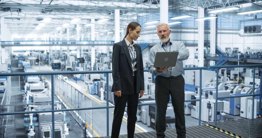 Portrait of a Male and Female Scientist Using Laptop Computer and Talking in a Factory Facility with Equipment Producing Modern Technical Devices for Different Industries