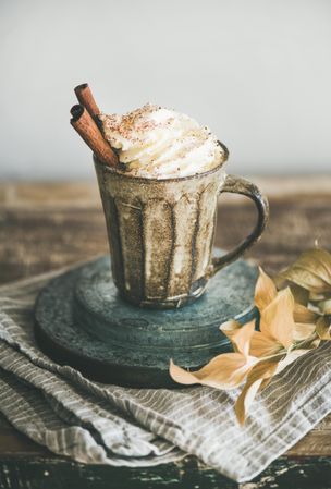 Mug of warm drink topped with whipped cream and cinnamon sticks, on light background, copy space