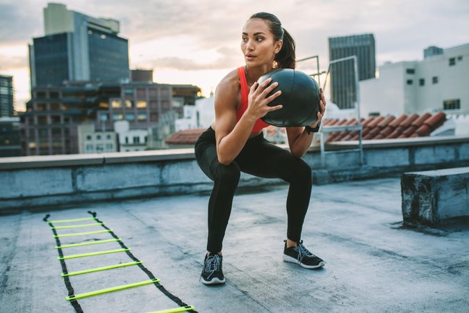 Woman doing squats holding a medicine ball with an agility ladder by her side on rooftop