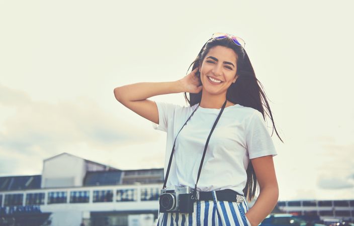 Woman in relaxed stance outside with camera around her neck and sunglasses on head