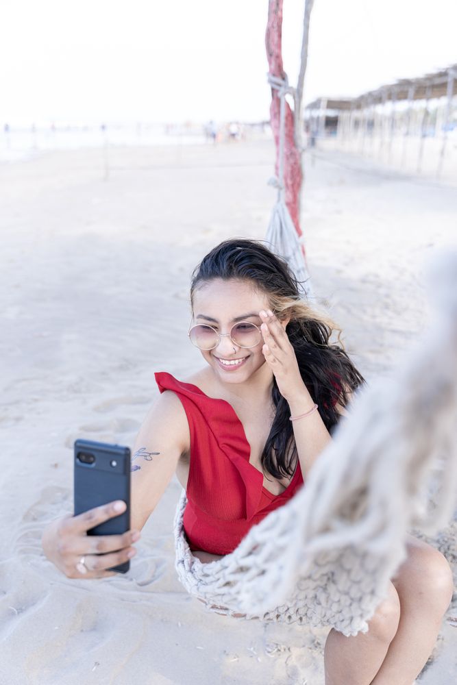A young Hispanic woman is smiling taking a selfie photo on a hammock at the beach