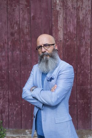 Front view of a mature trendy bearded hipster standing against a vintage red door outdoors while looking to camera