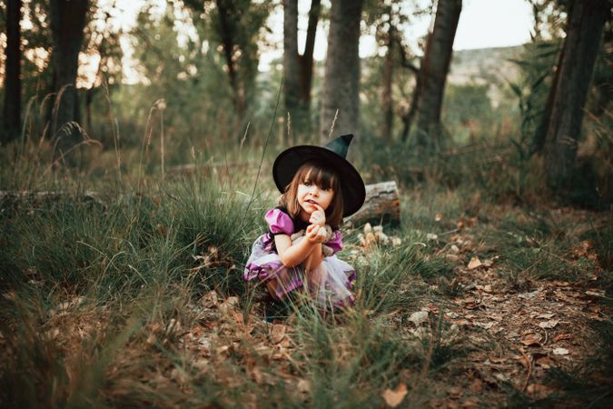 Girl in witch costume crouched down in the forest with finger to her mouth