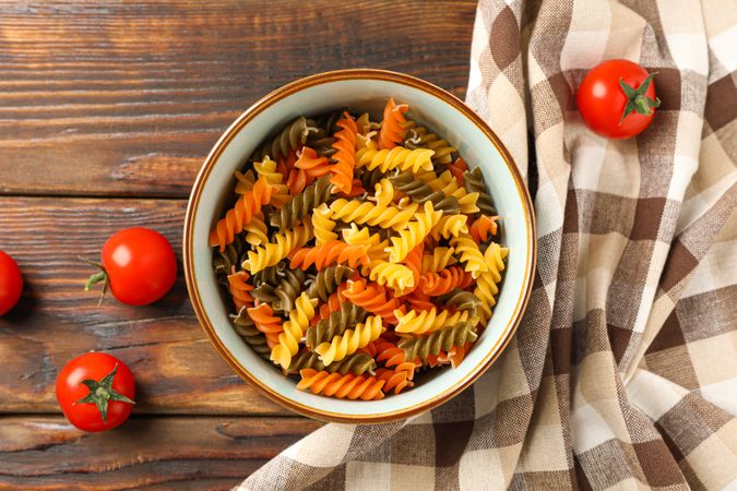 Bowl with color pasta, tomatoes and kitchen towel on wooden background, space for text