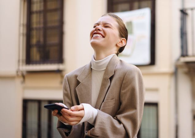 A Joyful and Young Woman Laughing While Enjoying Using Her Smartphone Outdoors in the Sun