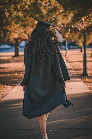 Back view of young woman in graduation gown walking on pathway between trees