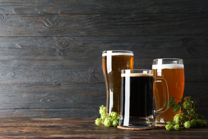Different mugs with beer and hop cones on wooden table, copy space