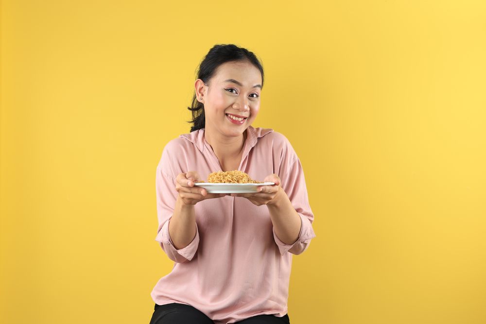 Asian Young Woman in Peach Shirt Holds a Plate of Mie Goreng Noodles with Happy Face.