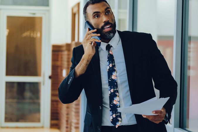Businessman in suit and tie with papers walking in office while speaking on smart phone