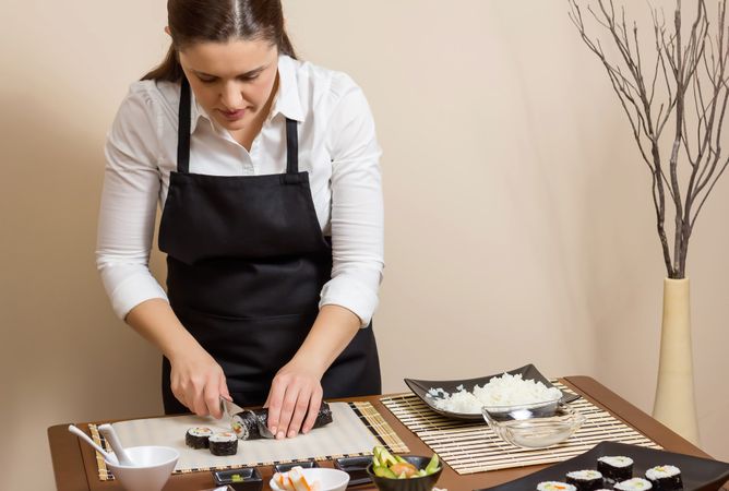 Female chef in apron cutting sushi rolls on table