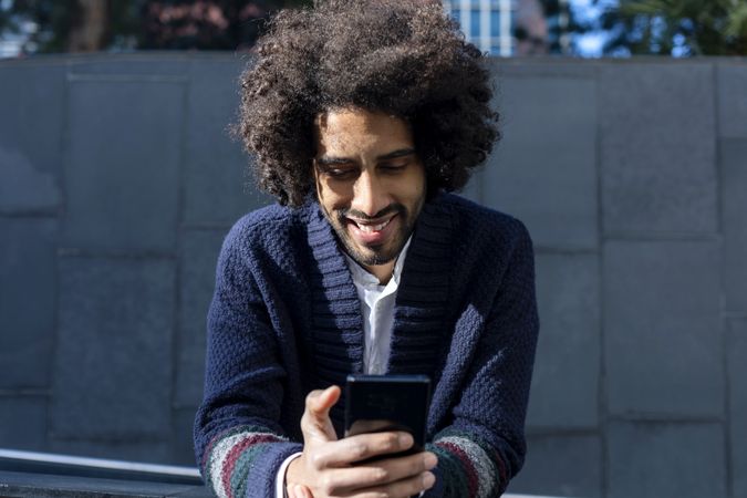 Black man in cardigan leaning forward outdoors on sunny day texting on phone
