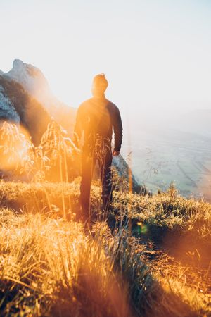 Back view of a man standing on brown grass facing the sun and mountains