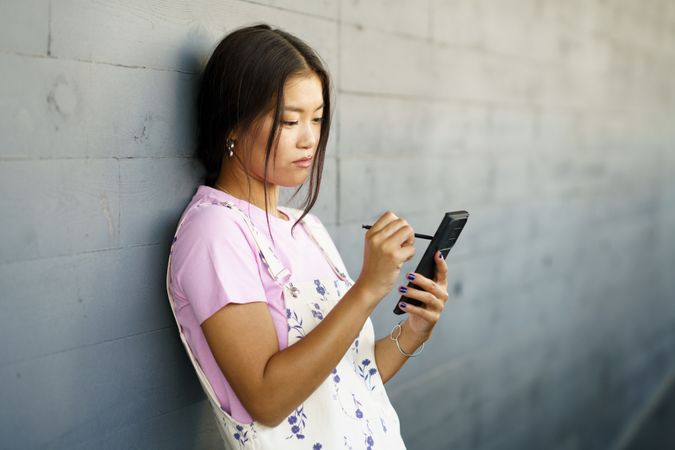Asian female wearing casual clothes holding phone while leaning against wall